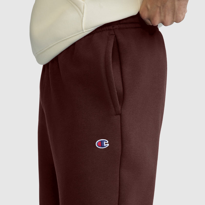 A person wearing maroon Champion Powerblend Sweatpants, Relaxed Bottom, C Logo and a cream-colored hoodie stands with one hand in their pocket. The iconic Champion C logo is visible on the left thigh, emphasizing the classic style of these men’s relaxed-bottom sweatpants.