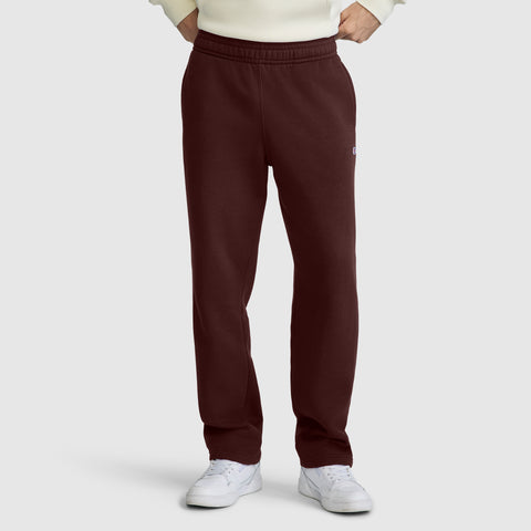 A person wearing maroon Champion Powerblend Sweatpants, Relaxed Bottom, C Logo, paired with a cream sweater and white sneakers, stands with hands in pockets against a plain light gray background.