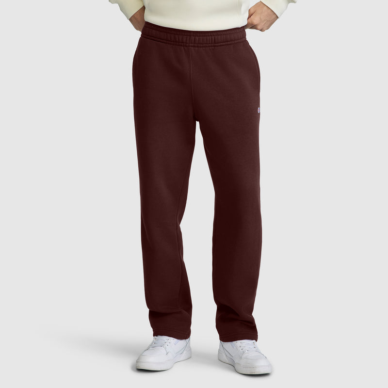 A person wearing maroon Champion Powerblend Sweatpants, Relaxed Bottom, C Logo, paired with a cream sweater and white sneakers, stands with hands in pockets against a plain light gray background.