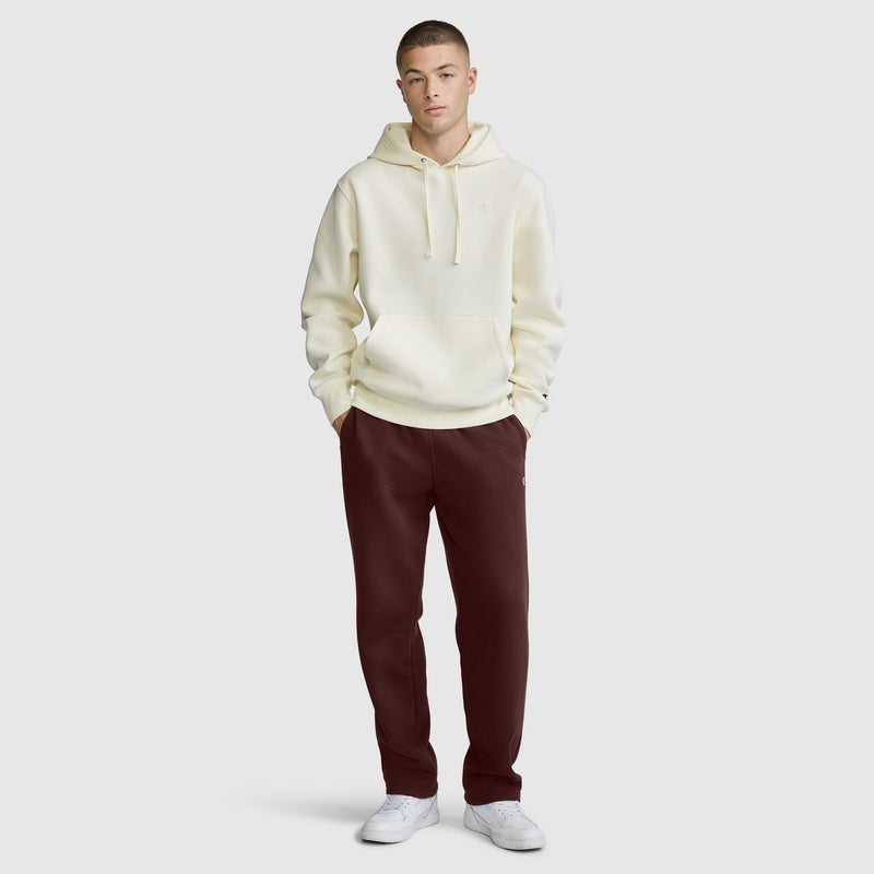 A person stands facing forward, wearing a cream hooded sweatshirt, Champion Powerblend Sweatpants with a relaxed bottom and C logo in dark maroon, and white sneakers against a plain light gray background.