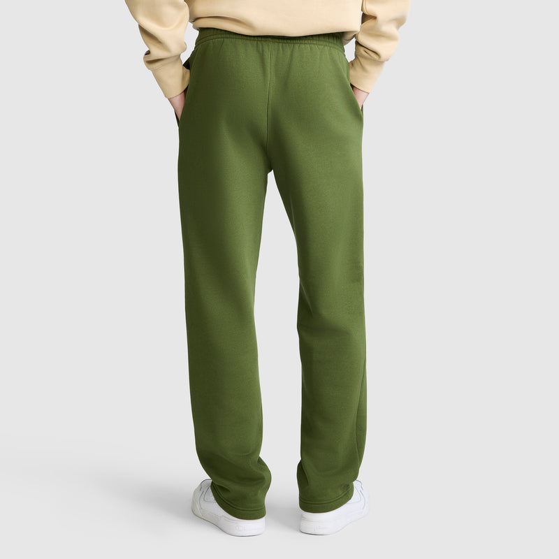 The person is wearing olive green Champion Powerblend Sweatpants, Relaxed Bottom, C Logo, made from a soft cotton blend. The sweatpants are paired with a beige sweatshirt and white sneakers. Standing with hands in pockets against a plain white background, only the lower half of the body is visible from the back.