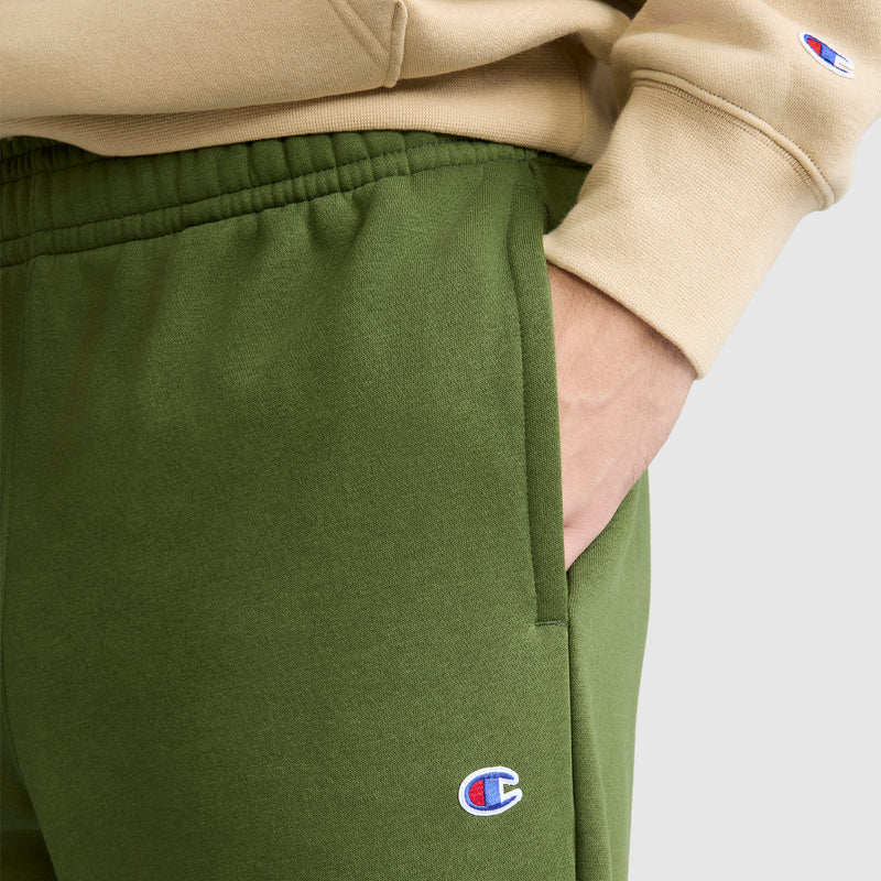 A person wearing green Champion Powerblend Sweatpants, Relaxed Bottom with the C Logo and a beige Champion sweatshirt stands with one hand in their pocket.