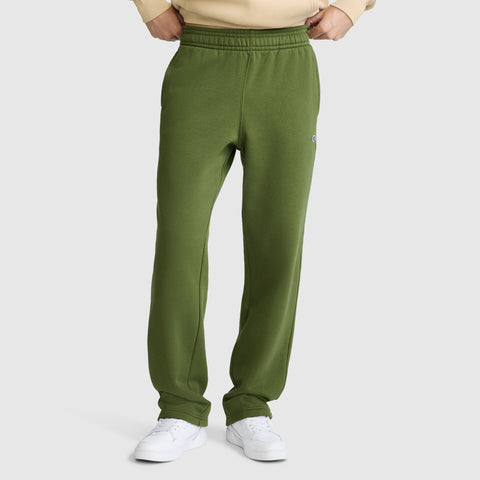 A person wearing green Champion Powerblend Sweatpants, Relaxed Bottom, C Logo and white sneakers stands with hands tucked into the waistband against a plain light background.