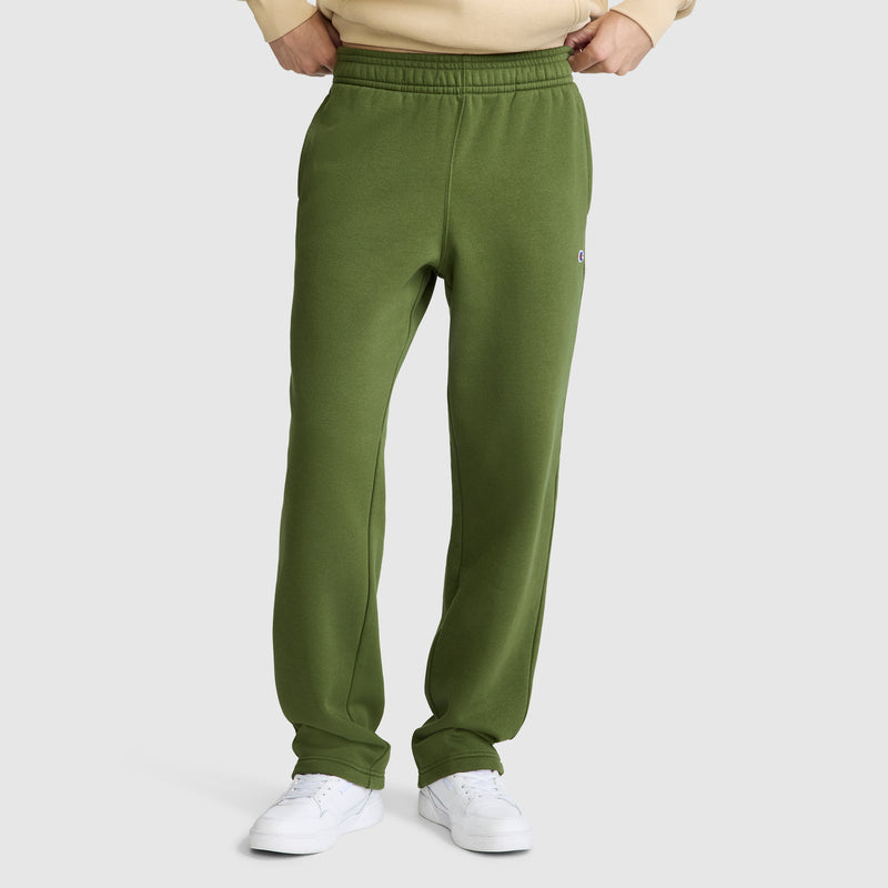A person wearing green Champion Powerblend Sweatpants, Relaxed Bottom, C Logo and white sneakers stands with hands tucked into the waistband against a plain light background.