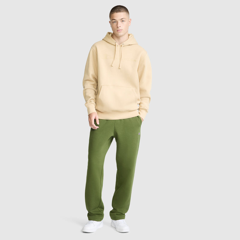 A young person stands against a plain light gray background, wearing a beige hoodie, Champion Powerblend Sweatpants with a relaxed bottom and C Logo in green—crafted from a soft cotton blend—and white sneakers.