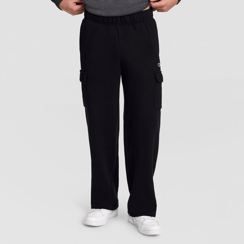 A person wears Champion’s Fleece Cargo Pant with a C Logo in black and pairs them with white sneakers, standing against a plain light gray background with their hands tucked into the waistband of the pants.