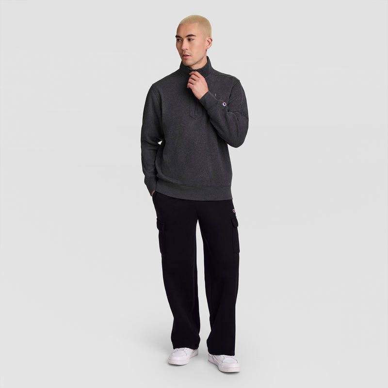 A person with short blond hair wears a dark gray pullover, Champion Fleece Cargo Pant, C Logo, and white sneakers, standing with one hand in their pocket against a plain light gray background.