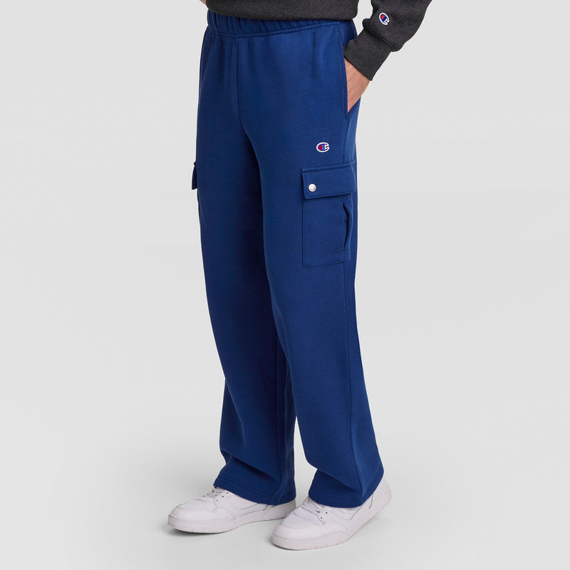 A person stands against a plain light background, one hand in their pocket, wearing Champion’s Fleece Cargo Pant, C Logo in blue. The men’s streetwear pants feature side flap pockets and a small logo, paired with white sneakers and a dark sweatshirt.