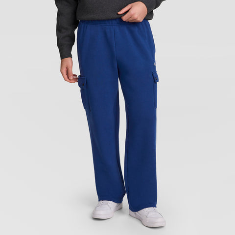 A person is wearing Champion’s Fleece Cargo Pant, C Logo in blue, paired with a dark gray sweatshirt and white sneakers, standing against a light gray background. Only the lower half of the body is visible, highlighting these stylish men’s streetwear pants from Champion.