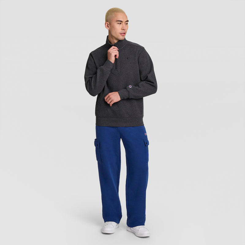 A person with short blond hair wears a dark gray quarter-zip sweater, Champion Fleece Cargo Pant with C Logo in blue, and white sneakers, standing against a plain white background and looking to the side.
