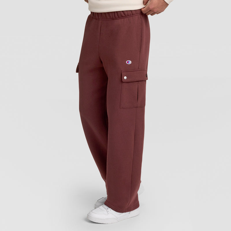A person wearing maroon Champion Fleece Cargo Pant, C Logo, featuring an elastic waistband and side flap pockets, paired with white sneakers. The visible Champion branding highlights the streetwear appeal of these pants against a plain light gray background.