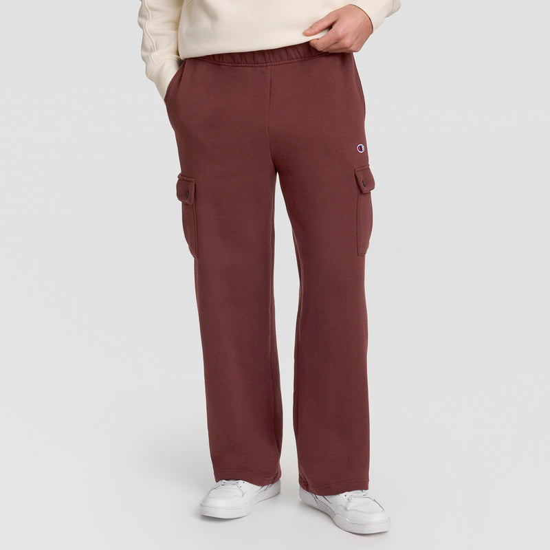 A person wearing Champion’s Fleece Cargo Pant with C Logo in maroon, featuring side pockets, styled in streetwear fashion with white sneakers and a beige sweatshirt, stands against a plain light background with hands in pockets.