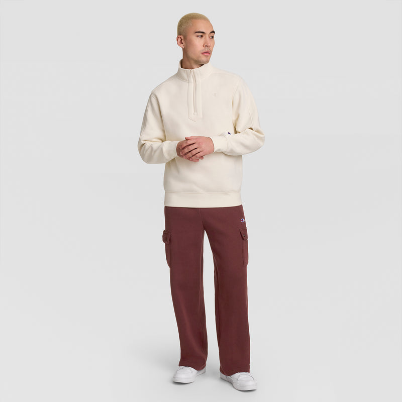 A person stands against a plain background, wearing a cream-colored pullover, white sneakers, and the Champion Fleece Cargo Pant, C Logo in maroon. With short blond hair and hands clasped while looking to the side, they effortlessly embody casual streetwear style.