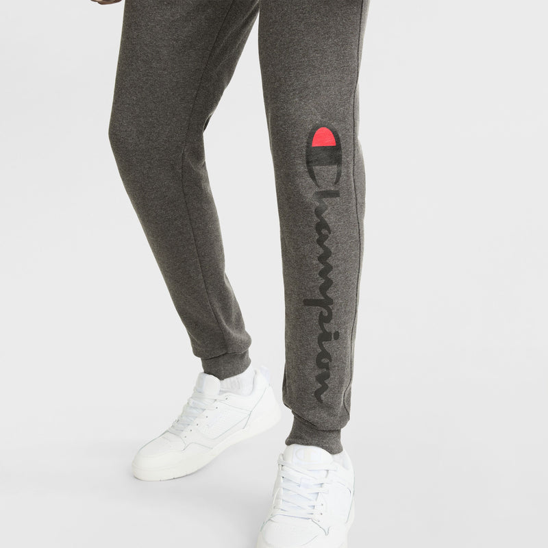 A person wearing dark gray Champion Powerblend Joggers featuring a script logo on the left leg and white sneakers, standing on a white background.
