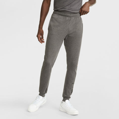 A person models Champion Powerblend Joggers, Script Logo, paired with a gray t-shirt and white sneakers, standing against a plain white background. The person's head is not shown in the image.