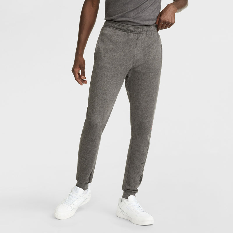 A person models Champion Powerblend Joggers, Script Logo, paired with a gray t-shirt and white sneakers, standing against a plain white background. The person's head is not shown in the image.