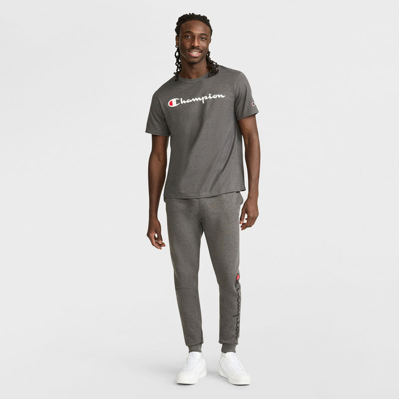 A man stands smiling in Champion Powerblend Joggers with Script Logo and white sneakers against a plain white background.