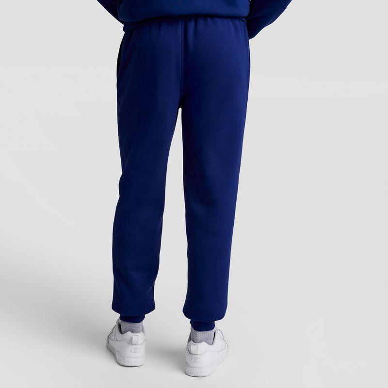 A person stands with their back to the camera against a plain light background, wearing dark blue Champion Powerblend Joggers with the Script Logo and white sneakers.