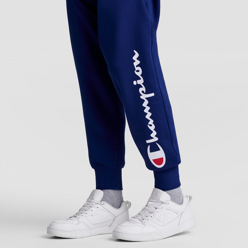 Someone stands against a plain light gray background wearing navy blue Champion Powerblend Joggers with the white script logo on the left leg, paired with white sneakers and light gray socks.