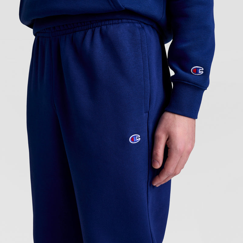 A person wears Champion Powerblend Joggers, Script Logo, and a matching navy blue sweatshirt with the Champion logo visible on both pants and left sleeve. Their right hand rests by their side against a plain background.