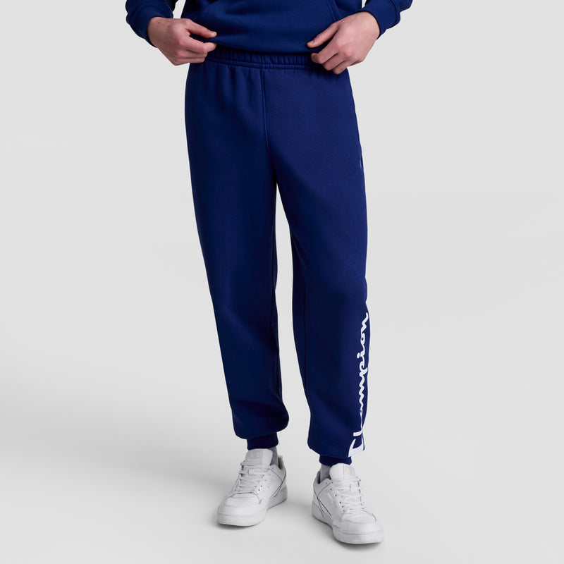 Someone dressed in Champion Powerblend Joggers with a white script logo on the left leg, a matching blue sweatshirt, and white sneakers stands against a plain light gray background.