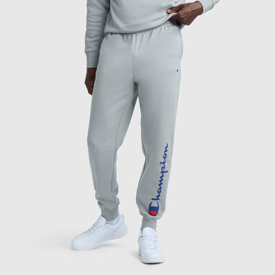 A person wears light gray Champion Powerblend Joggers with a blue and red script logo on the left leg, matching gray sweatshirt, and white sneakers, standing against a plain light background.