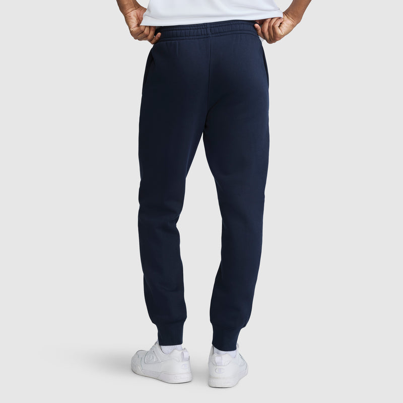 A person seen from the back with hands on their hips, wearing Champion Powerblend Joggers Script Logo in navy blue and white sneakers, standing against a plain light background.