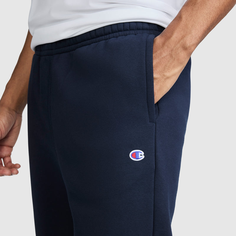 A person wearing Champion Powerblend Joggers with the script logo in navy blue, standing with one hand in the pocket against a plain light-colored background.