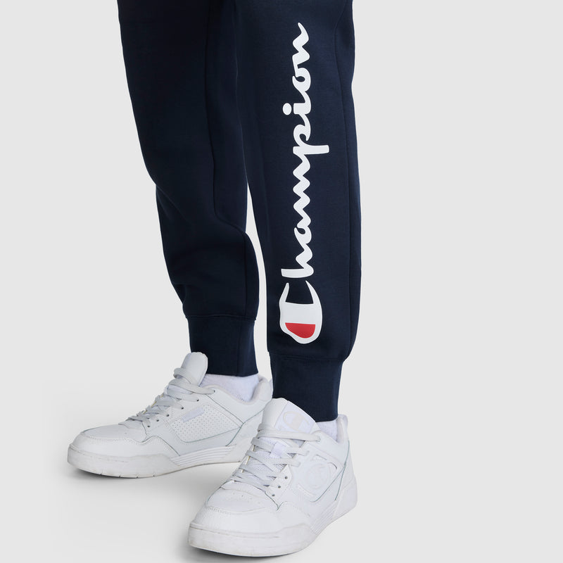 A person wearing navy Champion Powerblend Joggers with the white script Champion logo on the left leg, paired with white sneakers, standing on a plain light background.