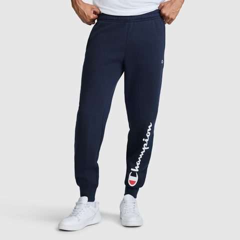A person wearing Champion Powerblend Joggers, Script Logo in navy blue with the white brand name on the left leg and white sneakers, standing against a plain light gray background.