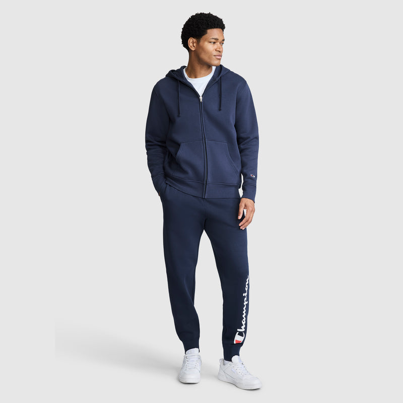 A man wearing a navy blue zip-up hoodie and Champion Powerblend Joggers with Script Logo, paired with white sneakers, stands facing slightly to the right against a light gray background.