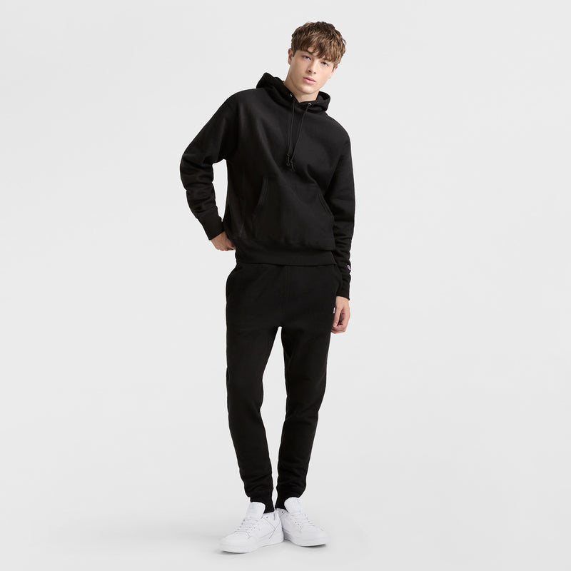 A young person stands against a white background, wearing Champion Reverse Weave Joggers with the C Logo, a black hoodie, and white sneakers, one hand in their pocket and the other on their hip.