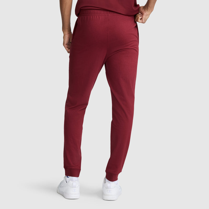 A person stands facing away, wearing Champion Lightweight Lounge Joggers with a C Logo in maroon and white sneakers. Their right hand rests on their hip as they pose against a plain light gray background.