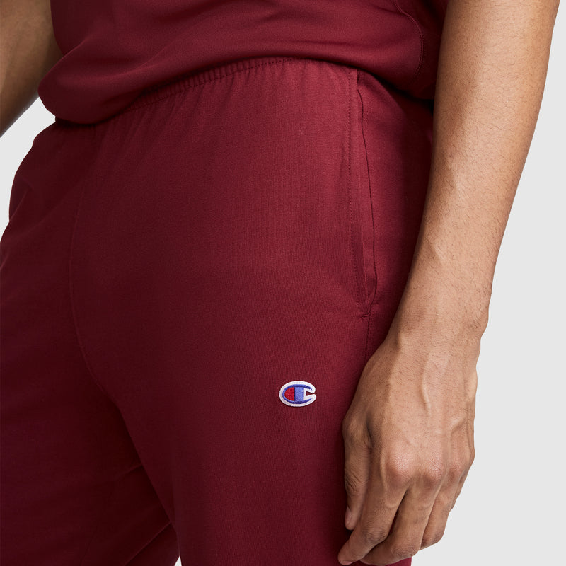A person is shown from the waist down, wearing Champion Lightweight Lounge Joggers in maroon, featuring a small C logo on the hip, posed against a plain light background with only their lower torso and right hand visible.
