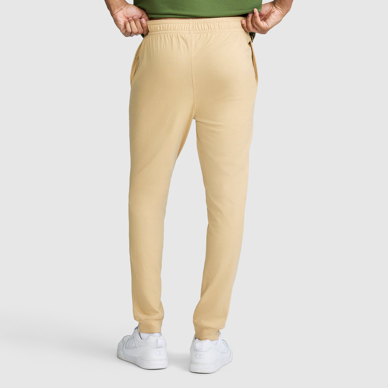 A person is shown from the back wearing Champion Lightweight Lounge Joggers with a C Logo in beige and white sneakers, hands tucked into the pockets. The top of a green shirt is partially visible.