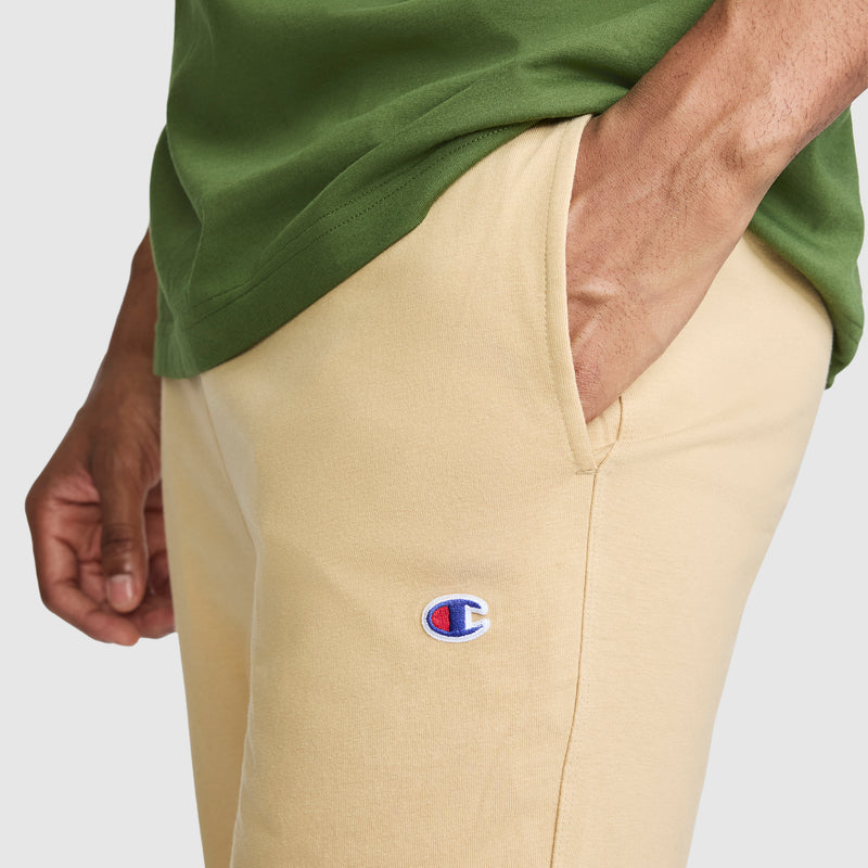 A person wearing a green shirt and beige Champion Lightweight Lounge Joggers, featuring the visible C logo, with one hand in the pocket.