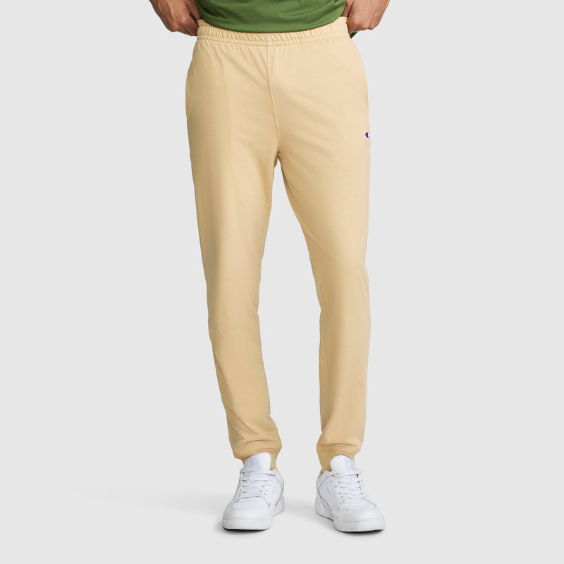 A person wearing Champion Lightweight Lounge Joggers with a C Logo in light yellow, paired with a green shirt and white sneakers, stands with hands in the waistband of the pants. The photo captures the outfit from the shoulders down against a light gray background.