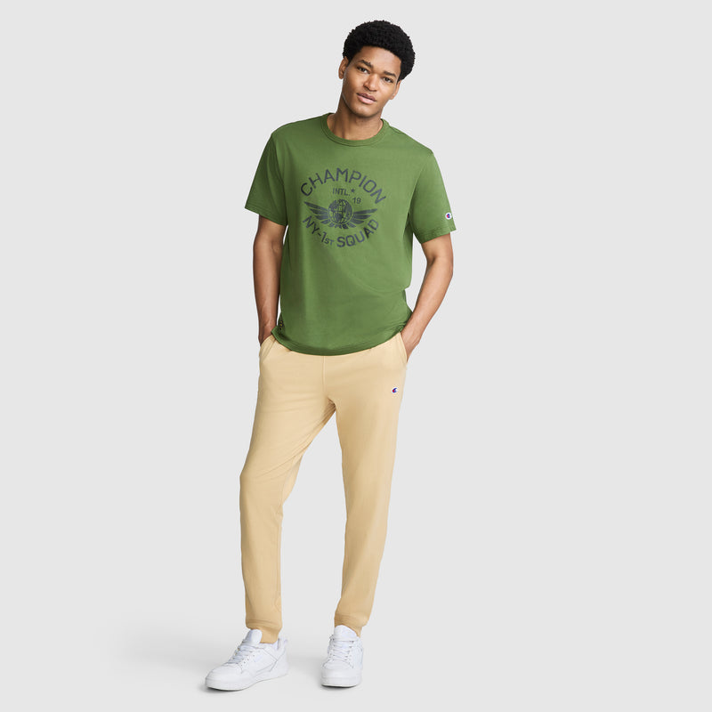 A man stands casually against a plain light background, wearing a green Champion t-shirt, Champion Lightweight Lounge Joggers with the C Logo in beige, and white sneakers.