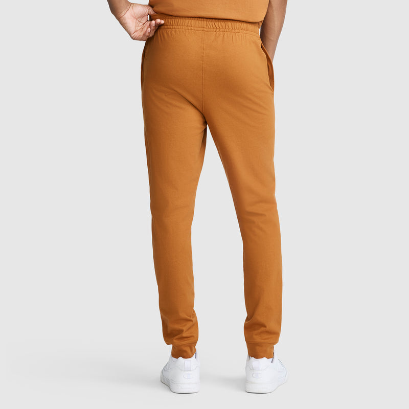 A person wearing Champion Lightweight Lounge Joggers, C Logo in orange and white sneakers stands facing away with one hand in a pocket, against a plain light gray background.