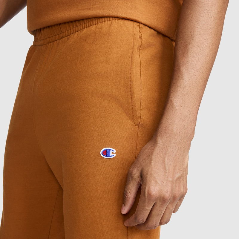 A person wearing orange Champion Lightweight Lounge Joggers, C Logo, with one hand relaxed by their side. The distinctive Champion logo appears on the upper thigh near the pocket, highlighting these men’s joggers as comfortable and perfect for everyday lounging.