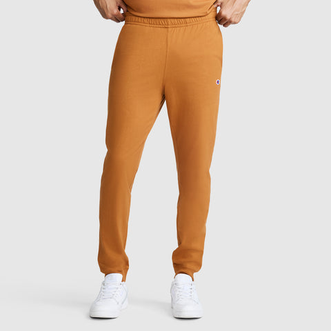 A person is wearing Champion Lightweight Lounge Joggers with a C Logo in orange, paired with white sneakers and an orange shirt, standing against a plain light gray background. Only the lower half of their body is visible.