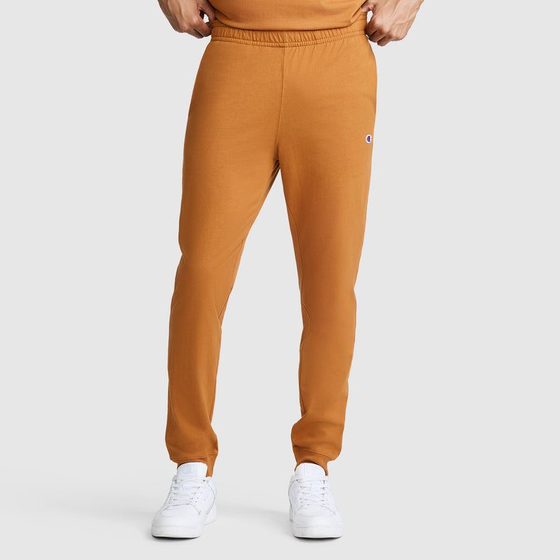 A person is wearing Champion Lightweight Lounge Joggers with a C Logo in orange, paired with white sneakers and an orange shirt, standing against a plain light gray background. Only the lower half of their body is visible.