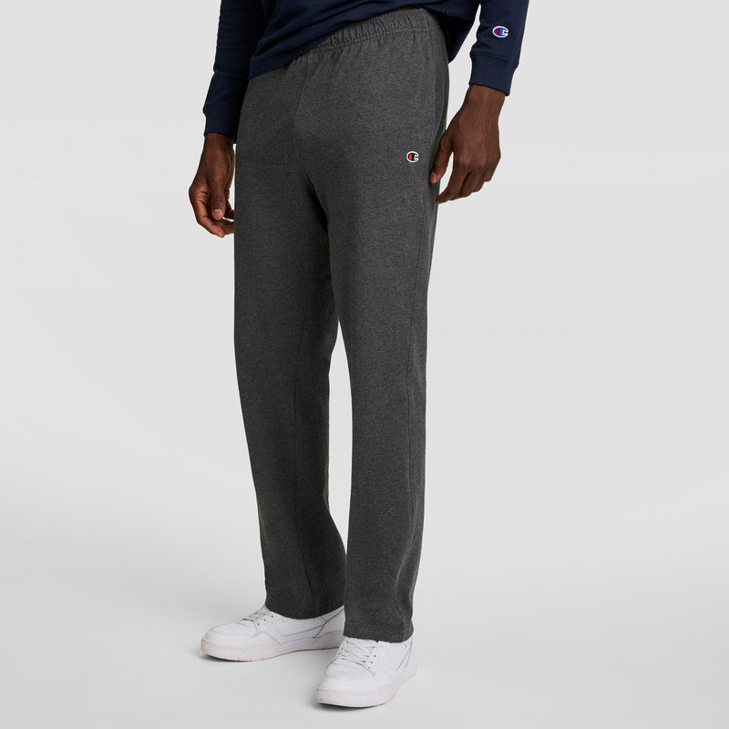 A person wearing dark gray Champion Lightweight Lounge Pants with an open hem and C logo, a navy long-sleeve shirt, and white sneakers stands against a plain light gray background.