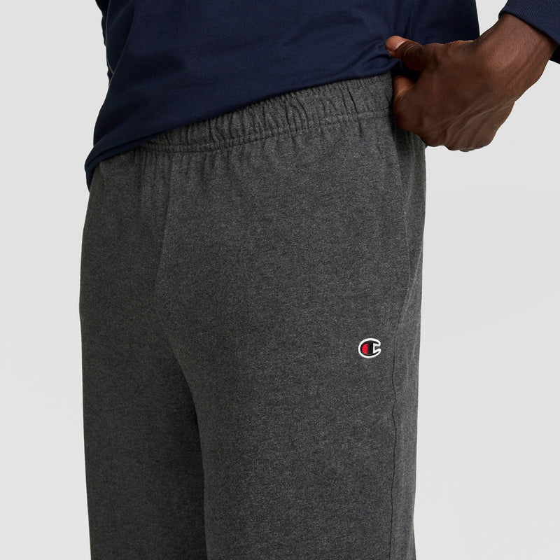 A person wears dark gray Champion Lightweight Lounge Pants with an open hem and C logo on the left thigh, paired with a navy blue shirt, and rests one hand on their hip.