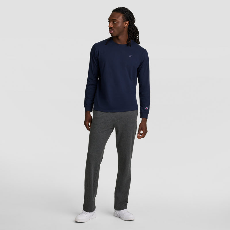 A man with long hair in twists wears a navy long-sleeve shirt, Champion Lightweight Lounge Pants with an open hem and C logo, and white sneakers while standing against a plain light gray background.