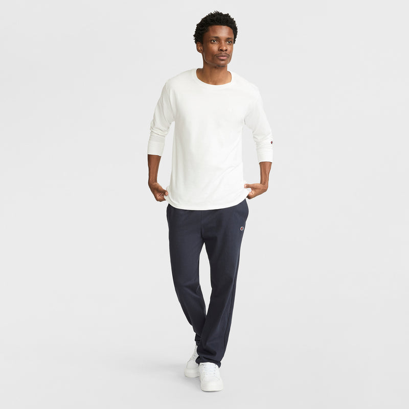 A man walks confidently in a long-sleeve white shirt, Champion Lightweight Lounge Pants with an open hem and C Logo for comfort, and white sneakers, set against a plain light gray background.