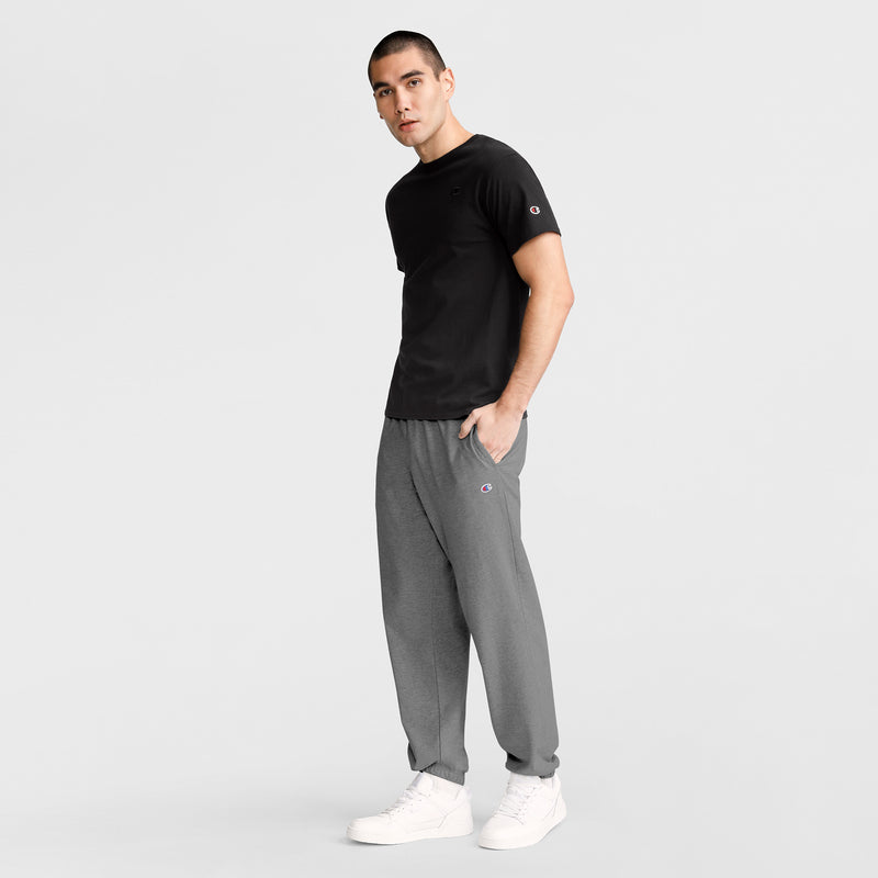 A man stands against a plain white background, wearing Champion Lightweight Lounge Pants with a cinched hem and C Logo, paired with a black T-shirt and white sneakers. One hand is in his pocket as he looks slightly to the side.