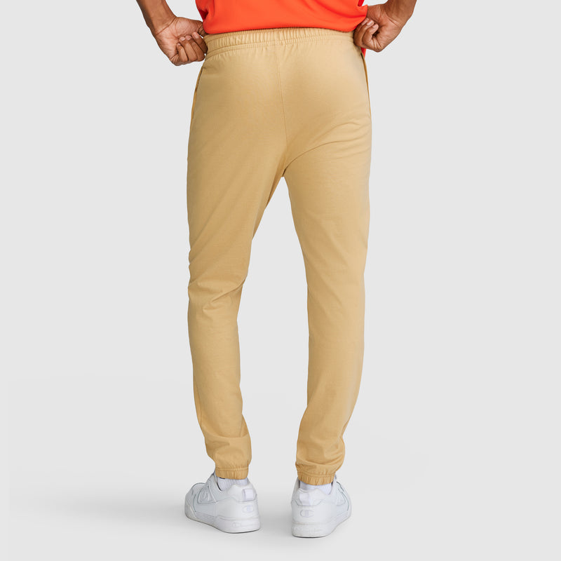 A person stands facing away, wearing Champion Closed Bottom Everyday Cotton Pants in tan—crafted from 100% cotton—paired with a tucked-in orange shirt and white sneakers against a light background.
