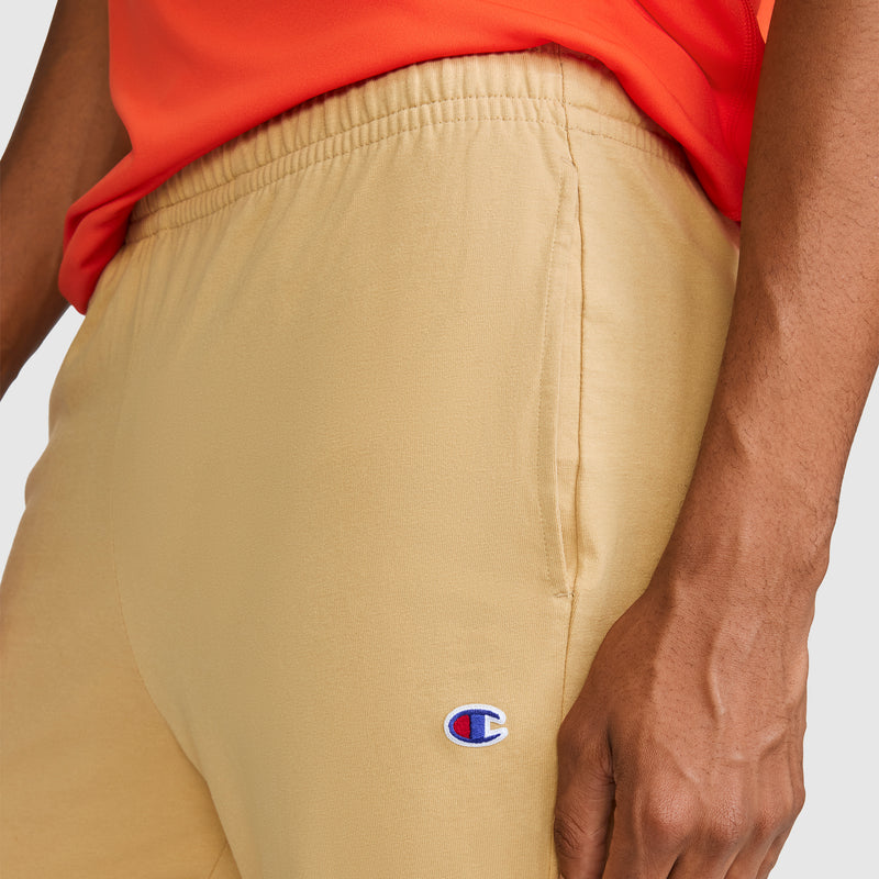 A person wearing Champion’s Closed Bottom Everyday Cotton Pant in tan and an orange shirt; only the lower torso and hand are visible.