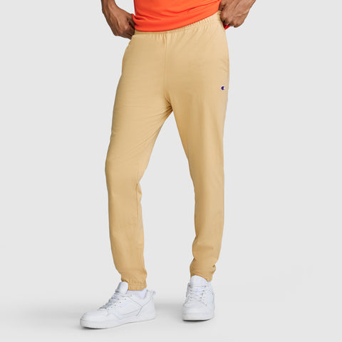 A person stands with hands resting on hips against a plain light-colored background, wearing Champion Closed Bottom Everyday Cotton Pants in beige crafted from 100% cotton, paired with a bright orange shirt and white sneakers.